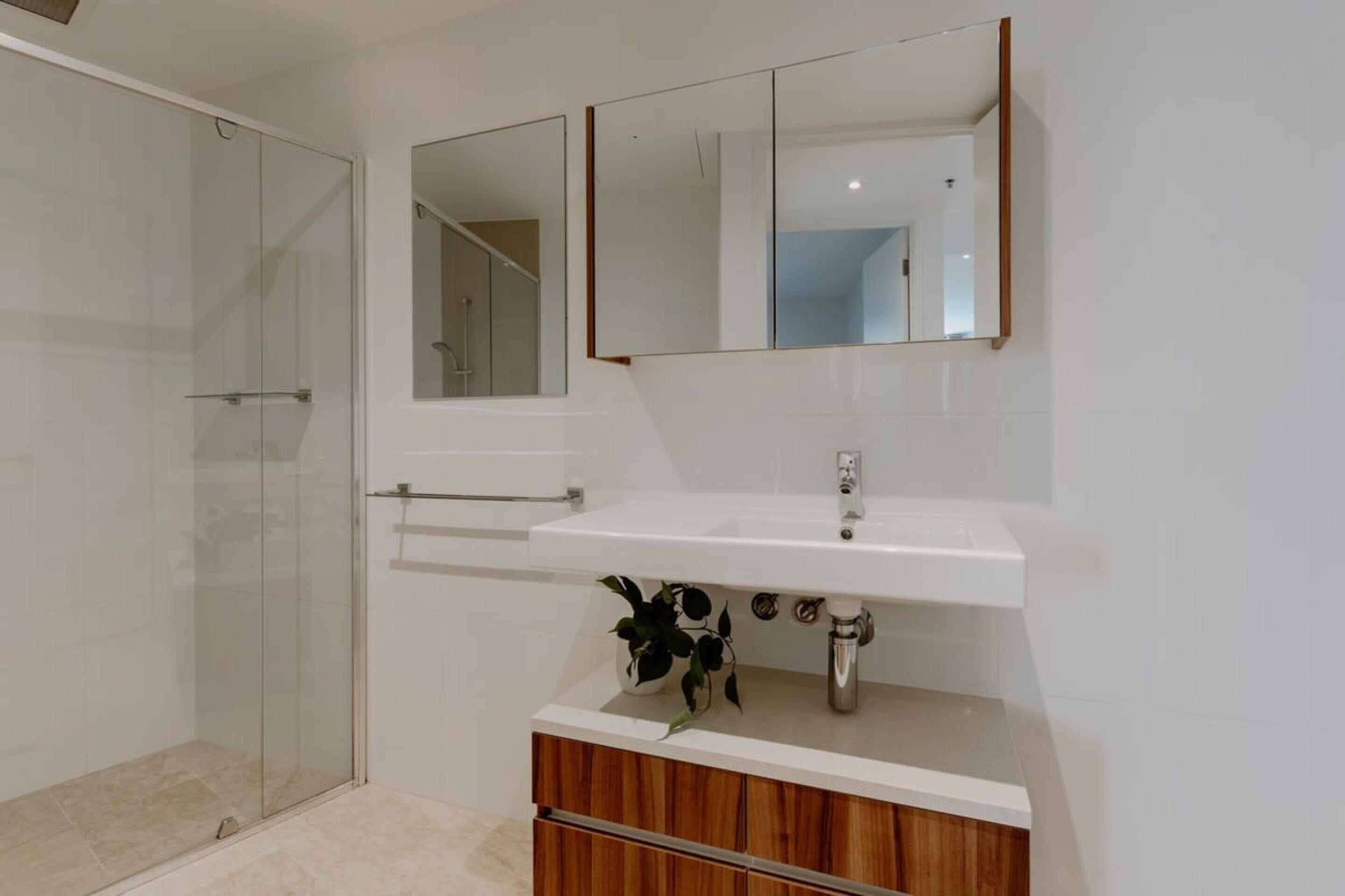 Une salle de bain avec une douche à parois de verre, un lavabo blanc avec un meuble-lavabo en bois, et un miroir au-dessus du lavabo.