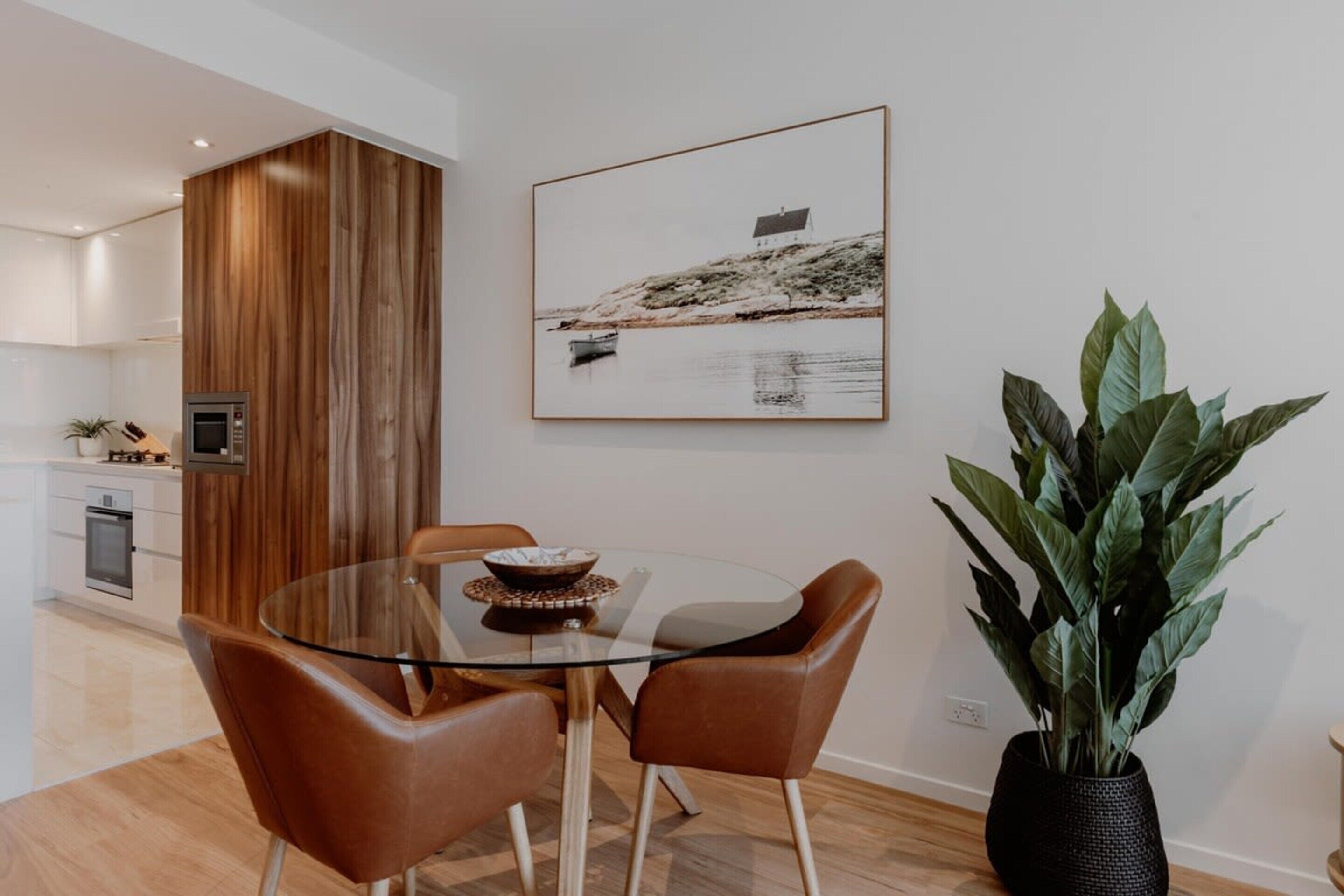 A modern dining area with a glass table, leather chairs, a wooden accent wall, a framed coastal landscape picture, and a large potted plant.
