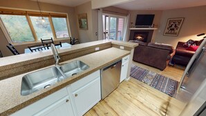 Eine moderne Küche mit Granit Arbeitsplatten, einer Edelstahlspüle und einem Geschirrspüler. Es gibt einen Essbereich mit Stühlen und Tisch, ein Wohnzimmer mit Sofa und Kamin sowie ein großes Fenster, das für natürliches Licht sorgt.