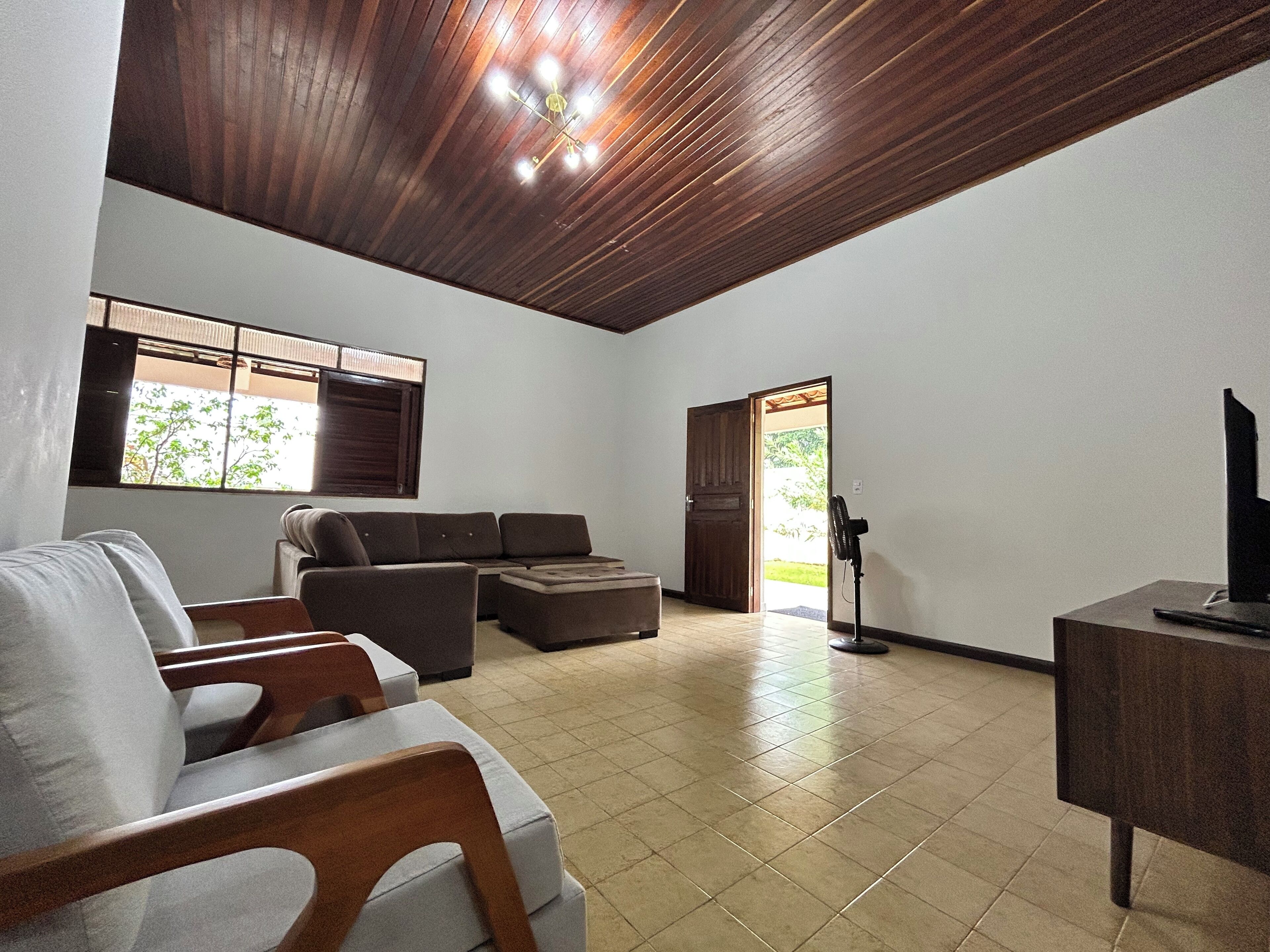 A spacious living area with a wooden ceiling, a large window with blinds, a sofa, a television on a stand, and a fan.