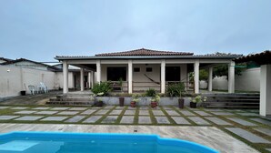 Ein Wohnhaus mit Schwimmbad, überdachter Terrasse und Gartenanlage mit Topfpflanzen.