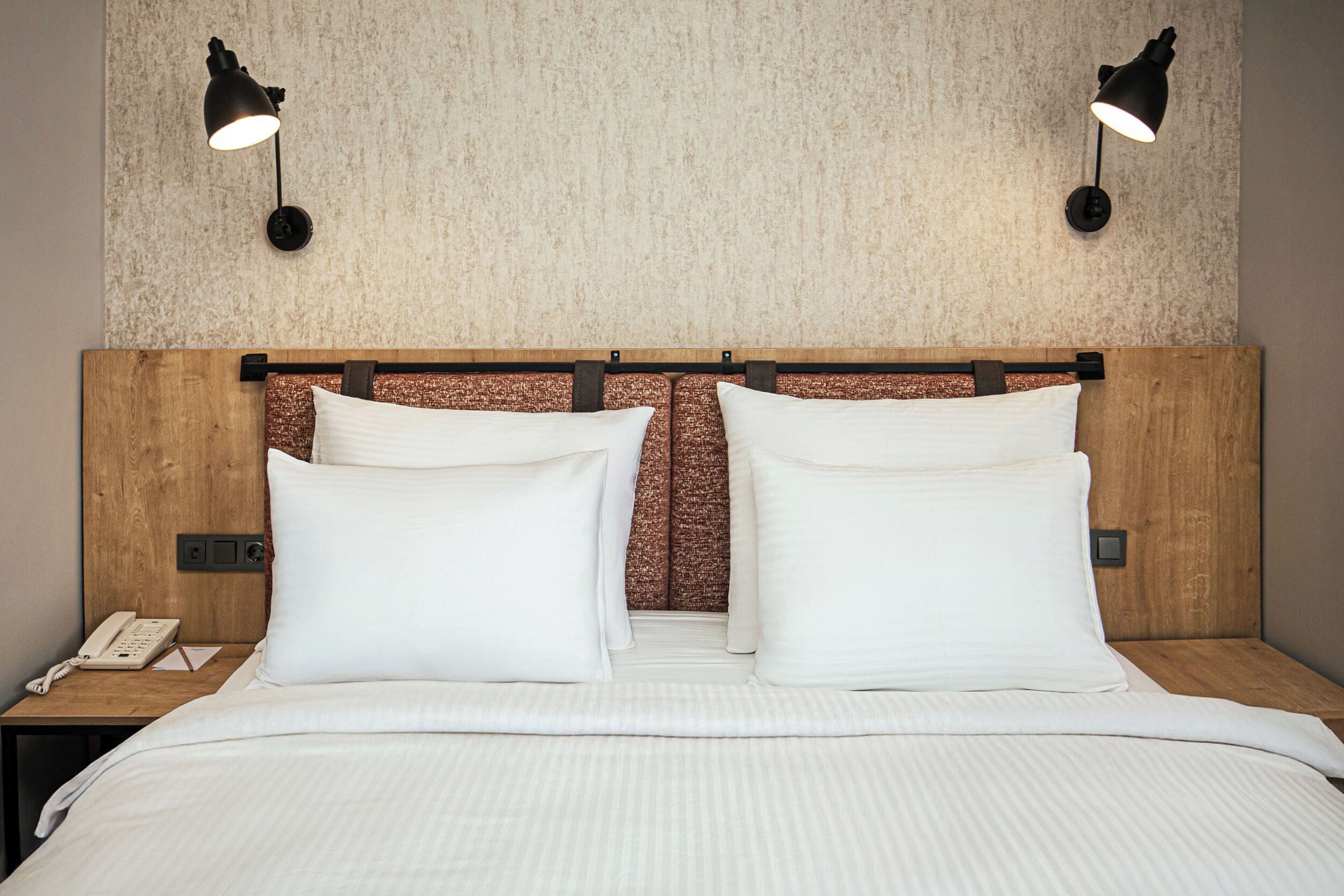 A neatly made bed with white linens, a wooden headboard, and two wall-mounted lamps.
