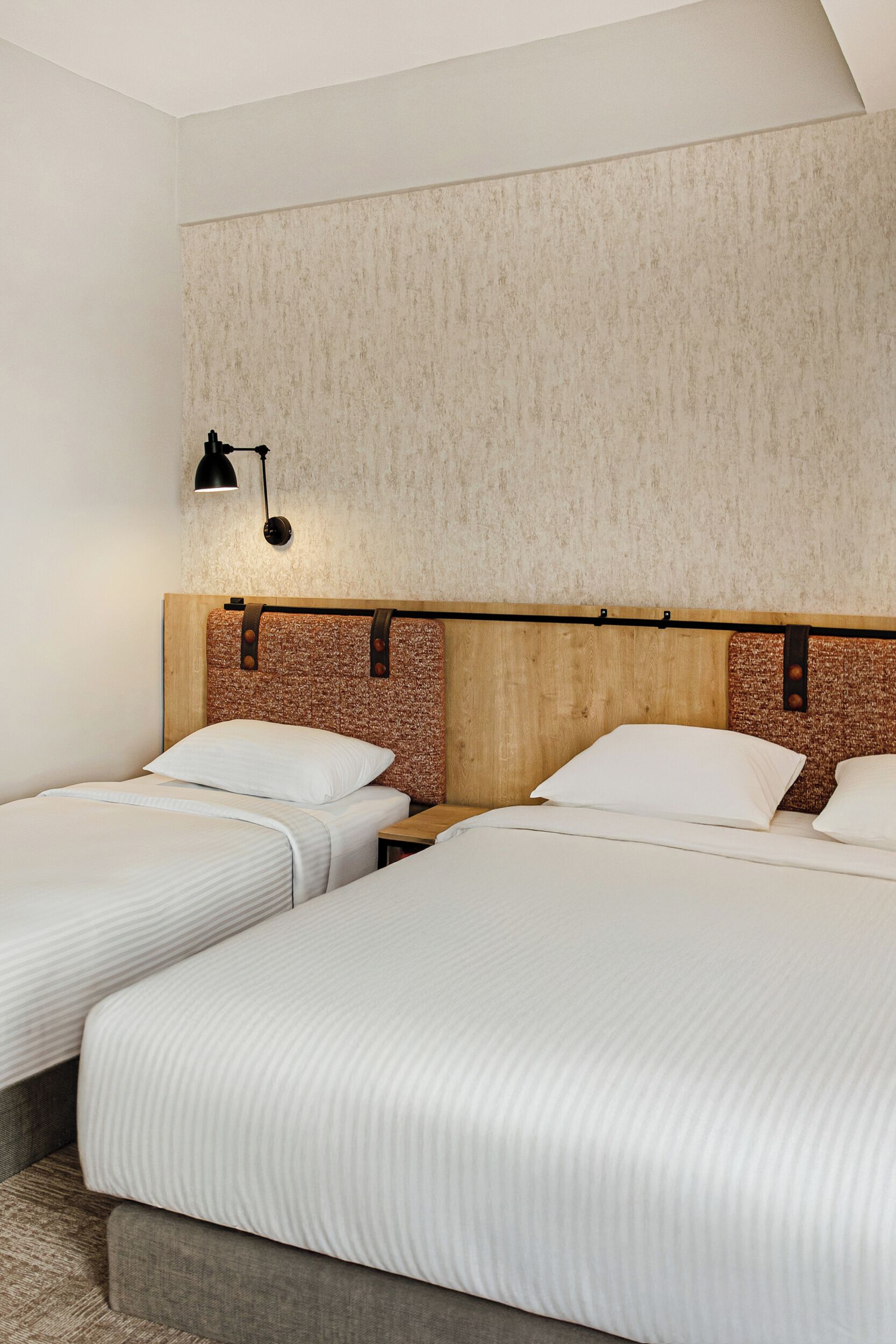 Une chambre d’hôtel avec deux lits simples, une tête de lit en bois, un mur à texture et une petite table de chevet avec une lampe.