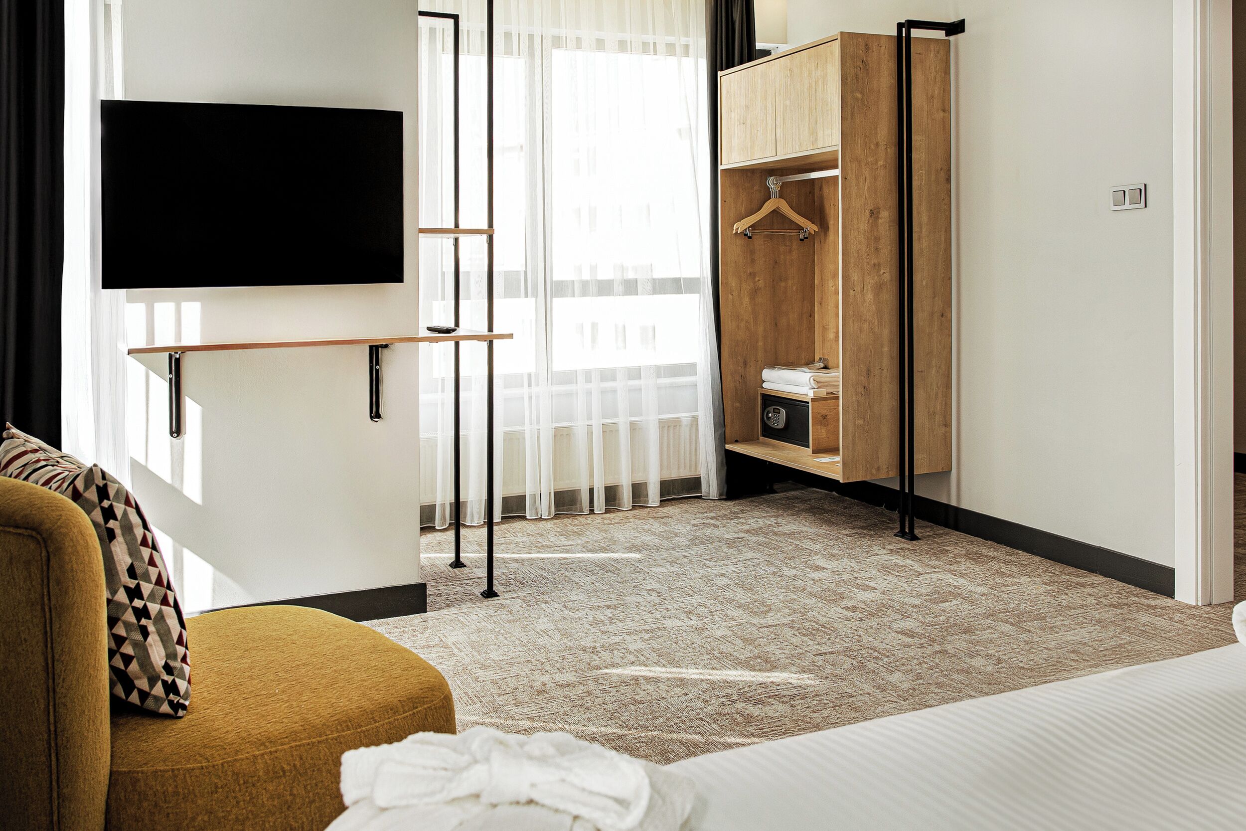Una habitación de hotel con cama, sofá, televisor montado en la pared, armario y ventana con cortinas de encaje.
