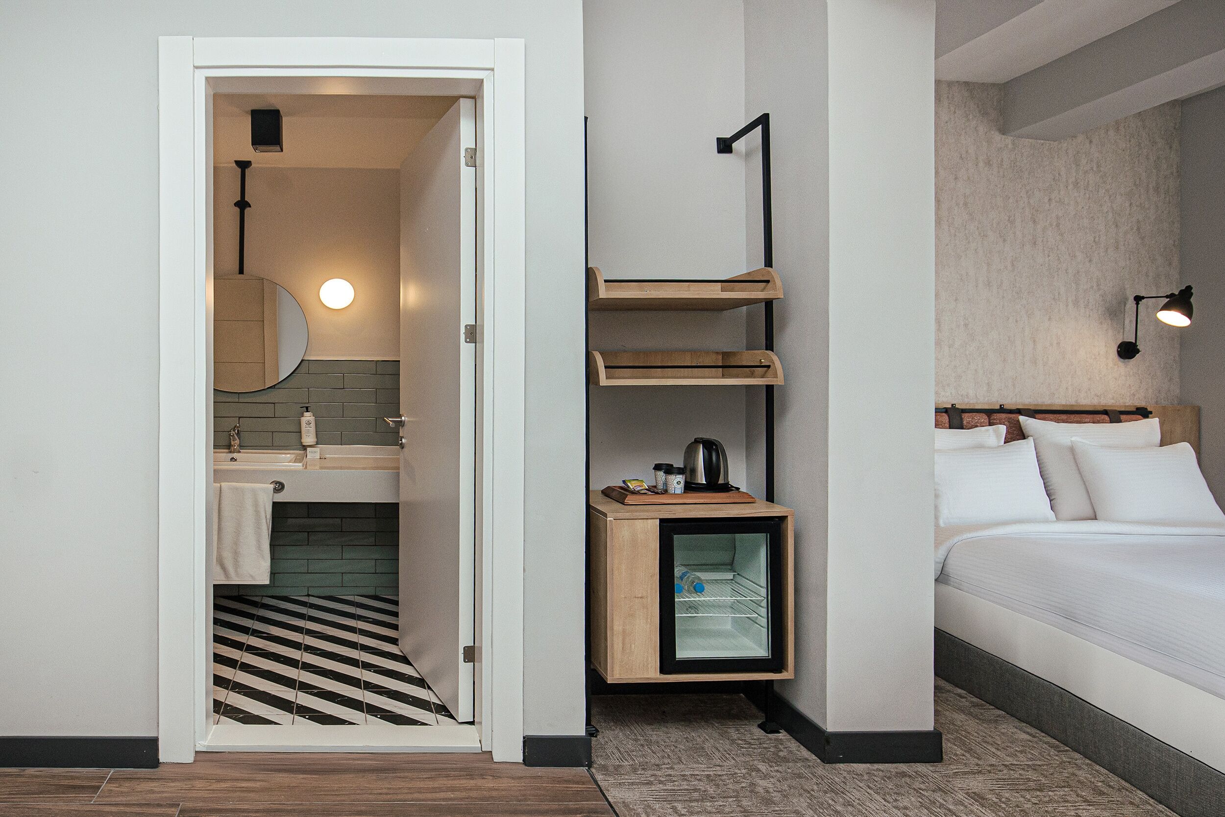 A modern hotel room with a bathroom, a bed, and a small wooden cabinet with shelves.