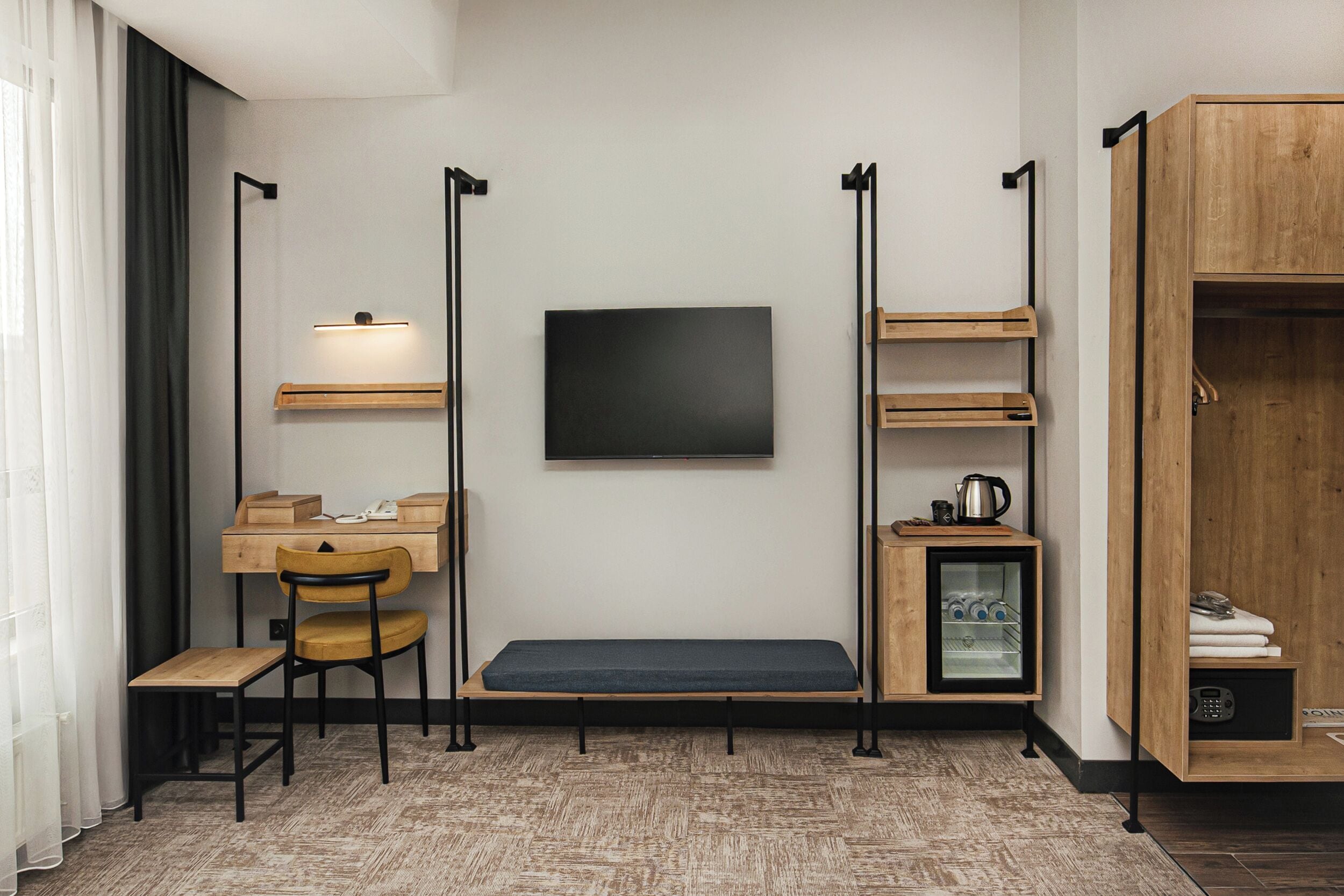 A compact room with a desk, chair, bench, TV, shelves, wardrobe, and a mini-fridge.