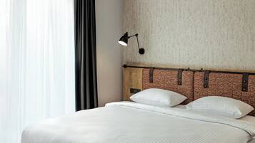Et moderne hotelværelse med en stor seng, en læderbeslået sengegavl, et natbord med lampe og udsigt til det frie gennem lette gardiner.