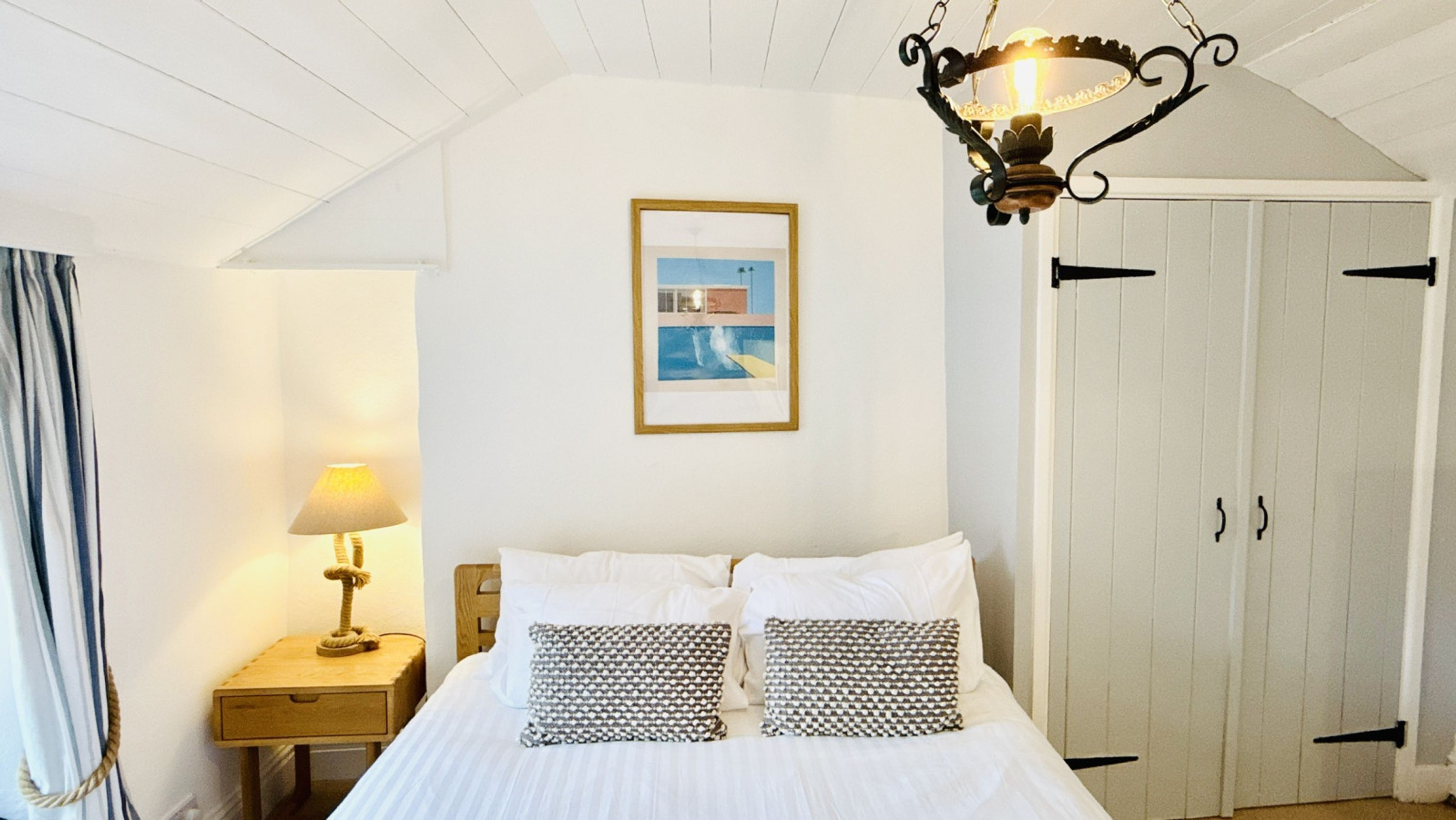 Et soveværelse med seng, et natbord med lampe, et billede i ramme på væggen og et garderobeskab.