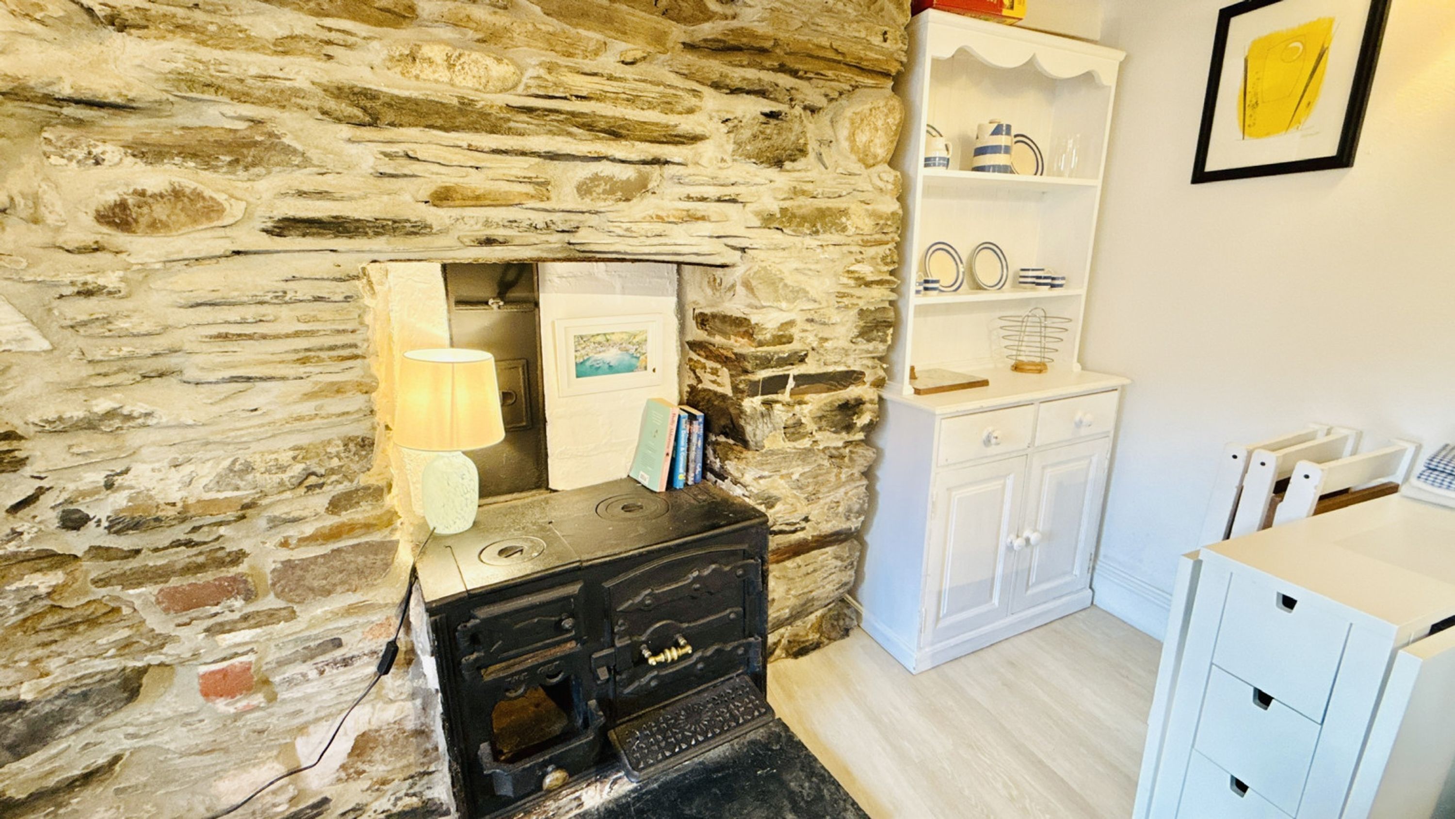 A rustic room with a stone wall, a vintage stove, a white cabinet, and a shelf with dishes.