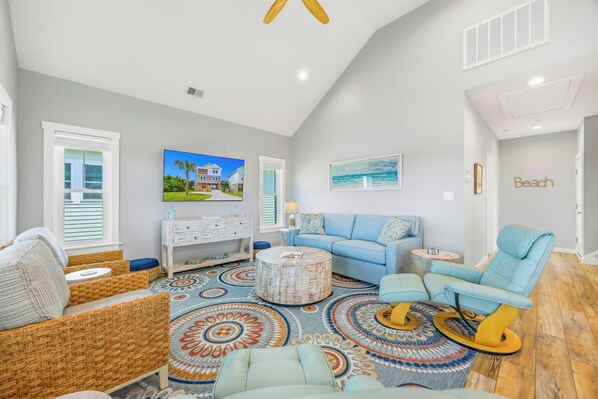 A spacious living room with a large window, a blue sofa, a wooden coffee table, and a patterned rug. - Brand New Soundside Avon Home with Hot Tub and Water Views (Avon)