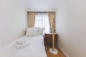 Una cama bien hecha con sábanas y almohadas blancas, una mesita de noche con lámpara, y una ventana con cortinas.