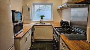 A compact kitchen with a microwave, stove, sink, and a window with a plant. - Ground Floor 1BDR Garden Flat South Croydon (Greater London)