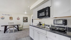 A kitchen with white cabinets, a black microwave, a toaster, and a sink. There is a chair and a table with a sign on the wall. - The Cozy Oak | Modern Basement Apartment (Fountain)