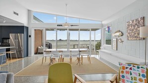 A modern living room with a large window overlooking a body of water, a kitchen with a stainless steel refrigerator, and a dining area with white chairs. - Riverside Retreat - Waterfront - Hindmarsh Island (Hindmarsh Island)