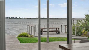 A view from a room with large windows overlooking a body of water, a wooden deck, and a grassy area with a bench. - Riverside Retreat - Waterfront - Hindmarsh Island (Hindmarsh Island)