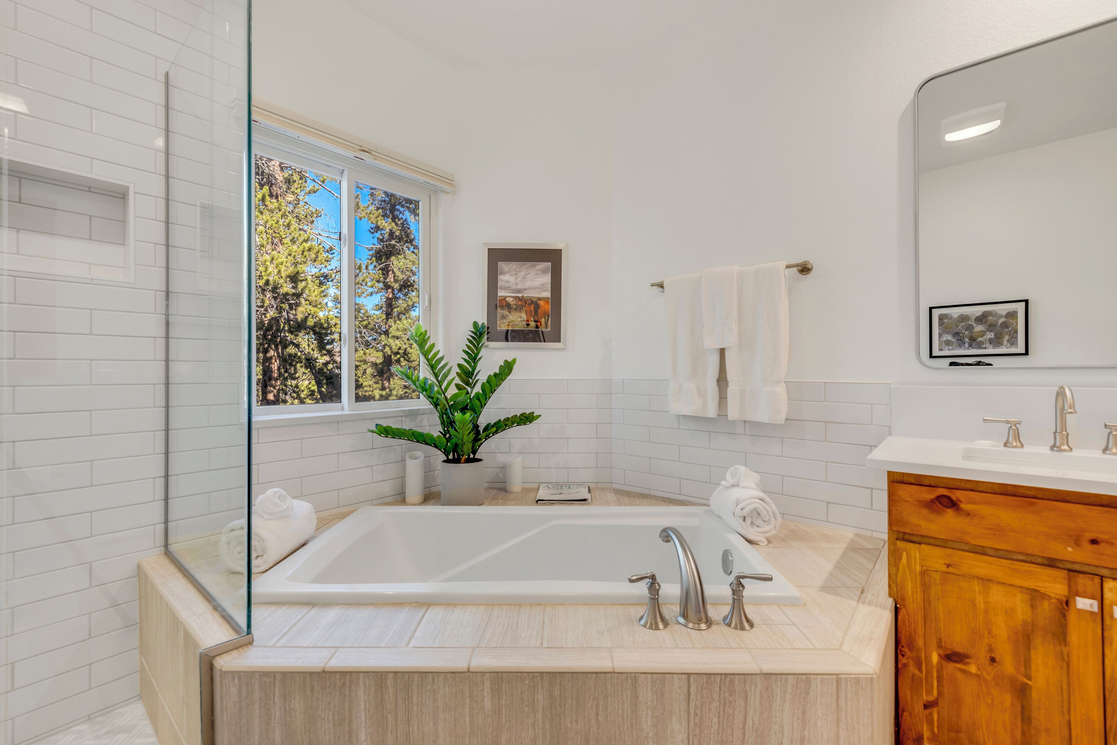 A modern bathroom with a large bathtub, a wooden vanity, a mirror, and a window with a view of trees.