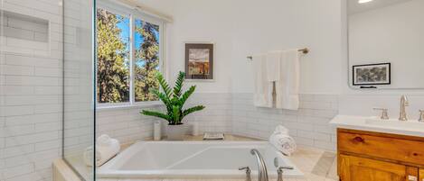 A modern bathroom with a large bathtub, a wooden vanity, a mirror, and a window with a view of trees.