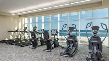 A gym with treadmills, ellipticals, and a stationary bike, featuring large windows with a view of the beach.