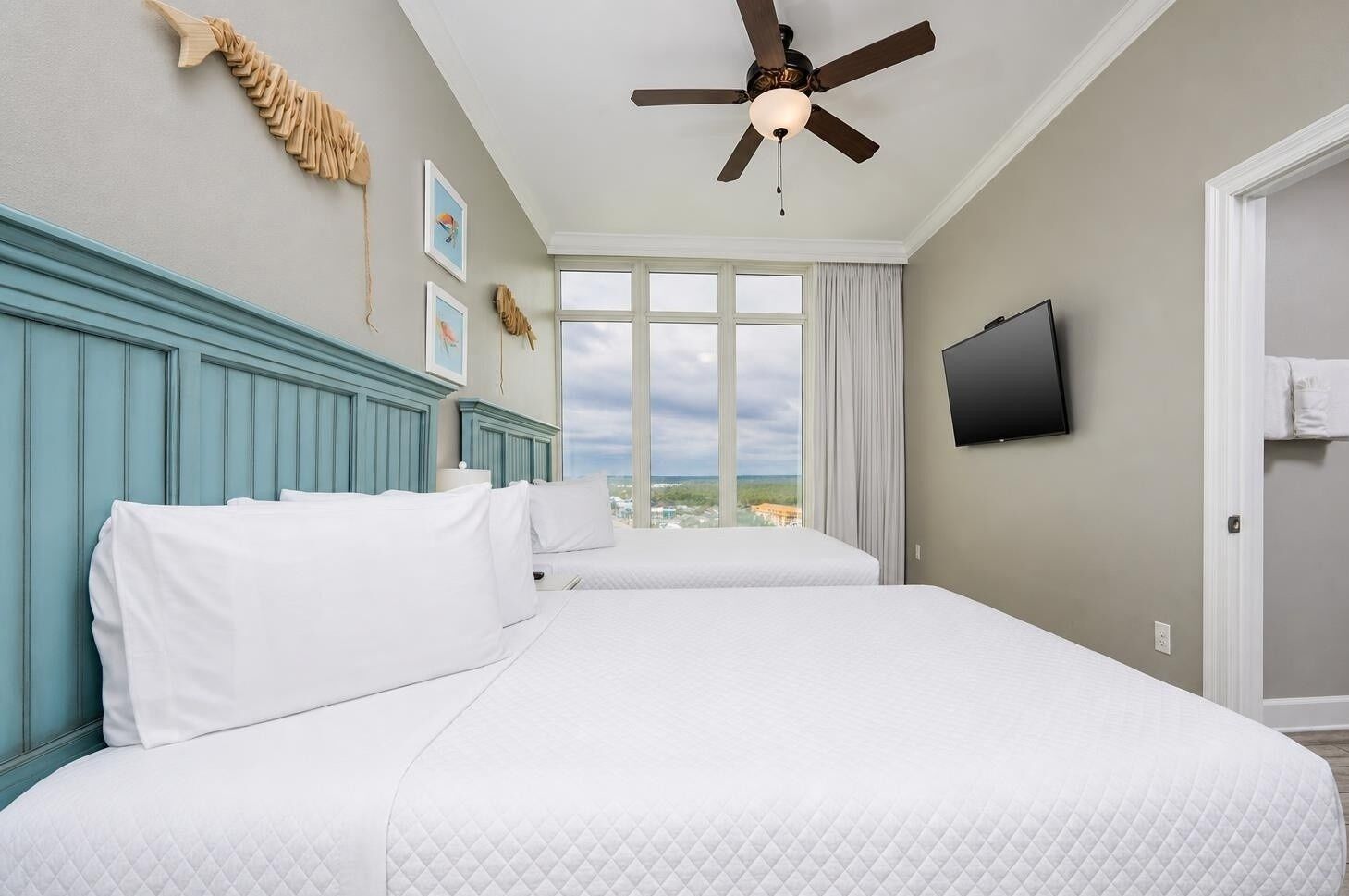 A hotel room with two beds, a ceiling fan, a TV, and a large window with a view.