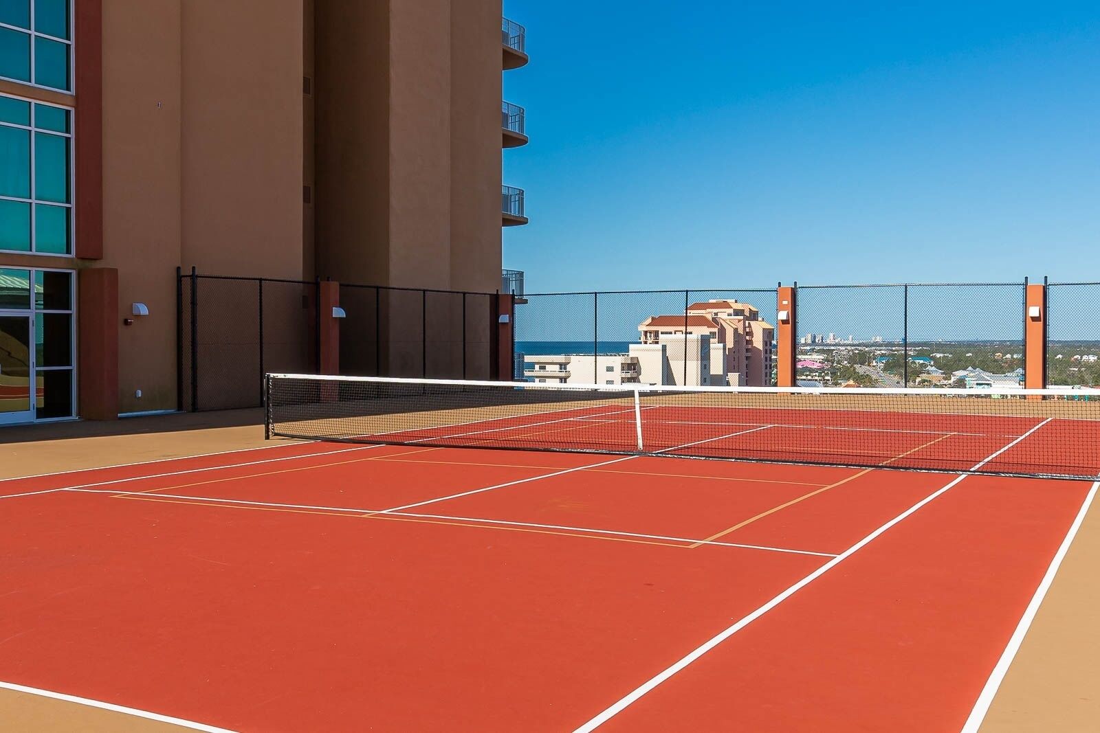 Un court de tennis en plein air, doté d’un filet et entouré d’une clôture, avec des bâtiments en arrière-plan.