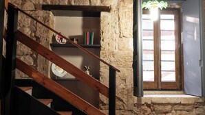 A staircase with wooden handrails leading to a stone wall. A shelf with books and a decorative plate is visible. A window with wooden shutters and a hanging light fixture is seen. - Monasteri Rural Holiday Home (Sicilia)
