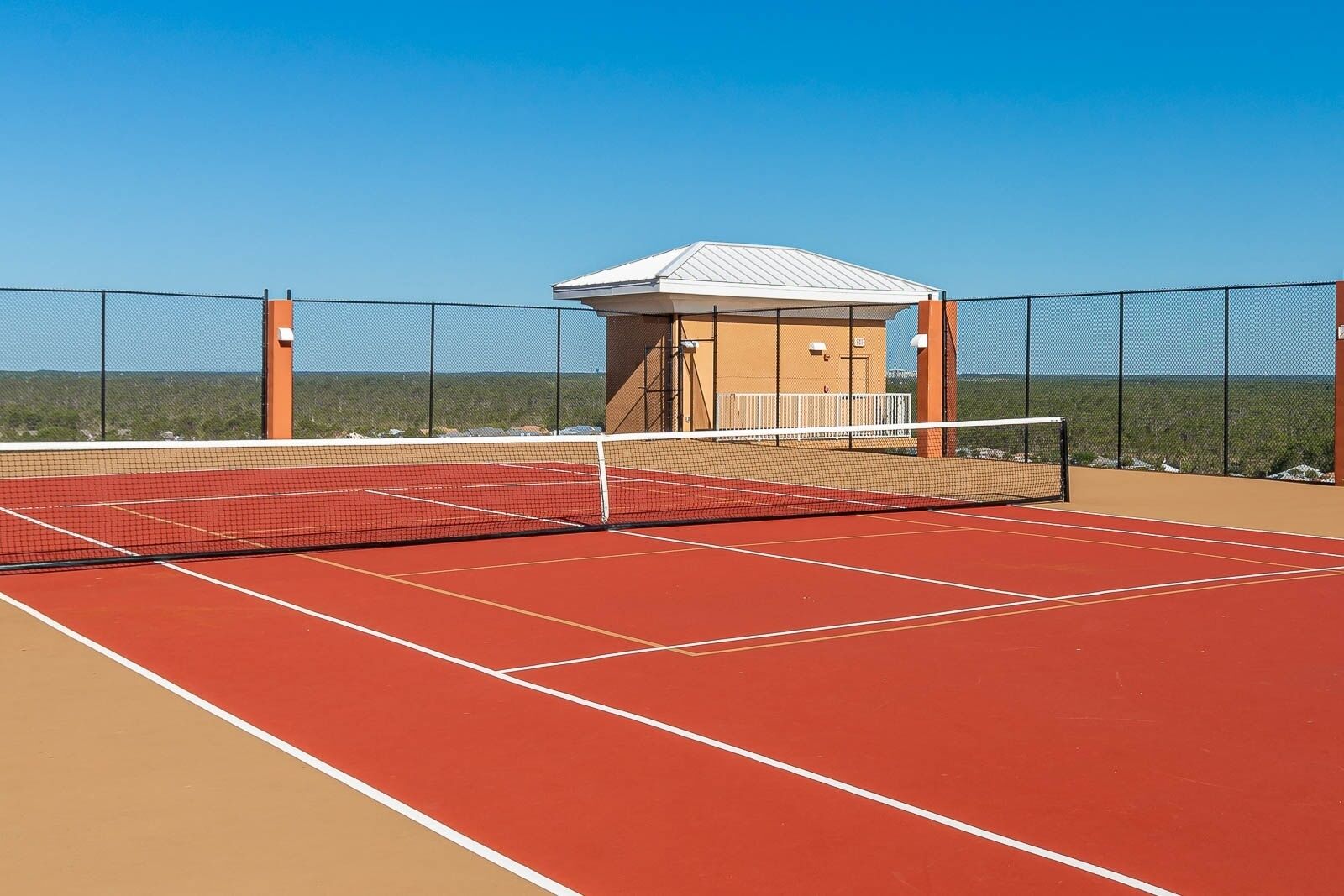 A tennis court with a net, surrounded by a fence and a small building with a white roof.