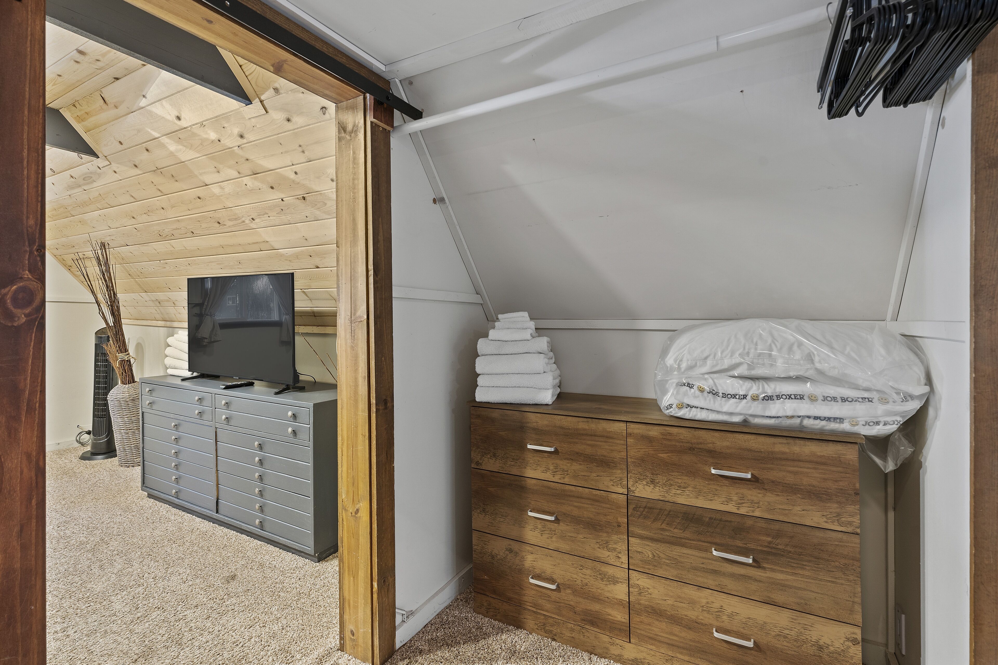 A room with a sloped ceiling, a grey dresser, a wooden cabinet, a television, and a stack of towels.