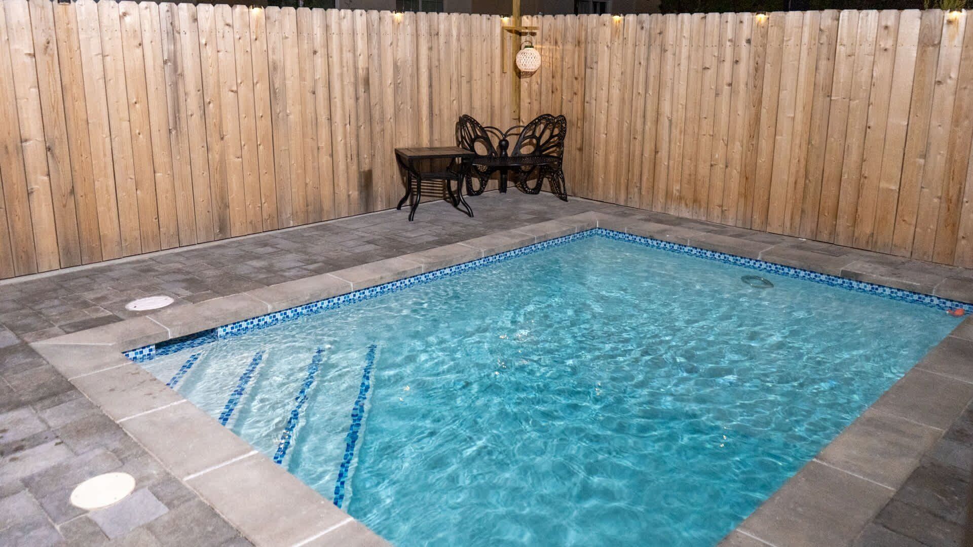 Piscina retangular interior com água azul límpida, rodeada por azulejos de pedra e uma cerca de madeira.