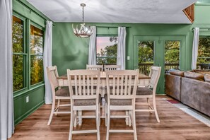 A dining area with a wooden table and chairs, a chandelier, and a view of the outdoors through large windows. - Smoky Mountain Bliss 2 Bedroom Cabin (Gatlinburg)