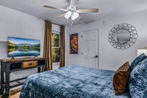 Ein Schlafzimmer mit einem Bett, einem Deckenventilator, einem Fernseher auf einem Schrank, einer Tür und einem an der Wand befestigten Spiegel.