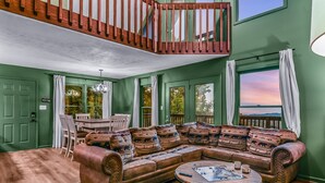 A green-walled living room with a brown leather sofa, a dining table, and a view of the sunset through a window. - Smoky Mountain Bliss 2 Bedroom Cabin (Gatlinburg)