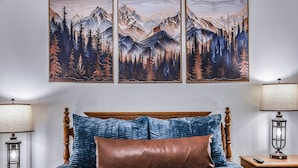 A bedroom with a wooden headboard, a bed with blue and brown bedding, two bedside lamps, and a wall-mounted painting of a forest and mountains. - Smoky Mountain Bliss 2 Bedroom Cabin (Gatlinburg)