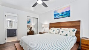 A bedroom with a bed, wooden headboard, bedside tables, a ceiling fan, a painting of the ocean, and a bathroom with a mirror and cabinets. - Big Island Fairway Terrace I-208 1 Bedroom Condo (Waikoloa)