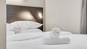 Een net opgemaakt bed met witte lakens en een opgerolde handdoek erop, een houten hoofdbord en een wandlamp.