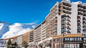 Un hôtel à plusieurs étages doté de balcons, un panneau indiquant la présence d’une station de ski, et des montagnes enneigées en arrière-plan.