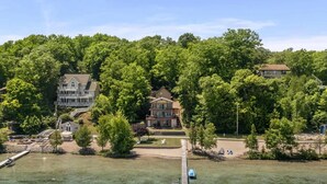 Una proprietà sul lago con un molo, una casa grande circondata da alberi e un edificio più piccolo.