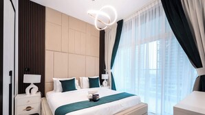 Una habitación de hotel moderna con una cama grande, dos mesitas de noche con lámparas, vista a la ciudad a través de cortinas translúcidas y un paisaje urbano visible desde la ventana.