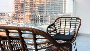 Una silla de mimbre con un cojín negro en un balcón con vistas a un paisaje urbano y edificios altos.
