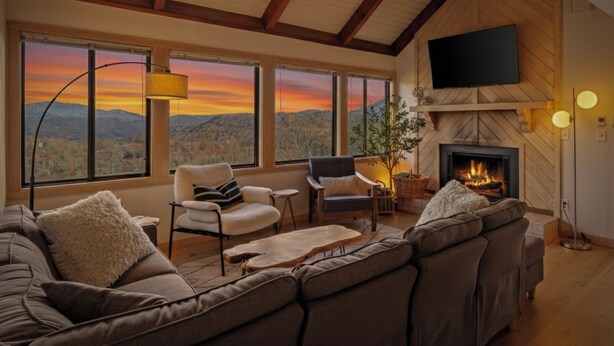 A cozy living room with a fireplace, large windows with a mountain view, and a ceiling fan. - “High Haven — A Modern Mountain Retreat with Breathtaking Views” (Sugar Mountain)