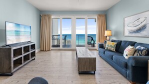 A living room with a TV, a wooden coffee table, a blue sofa, and a view of the ocean through large windows. - Waterfront condo w/ gulf views & resort pools (Panama City Beach)