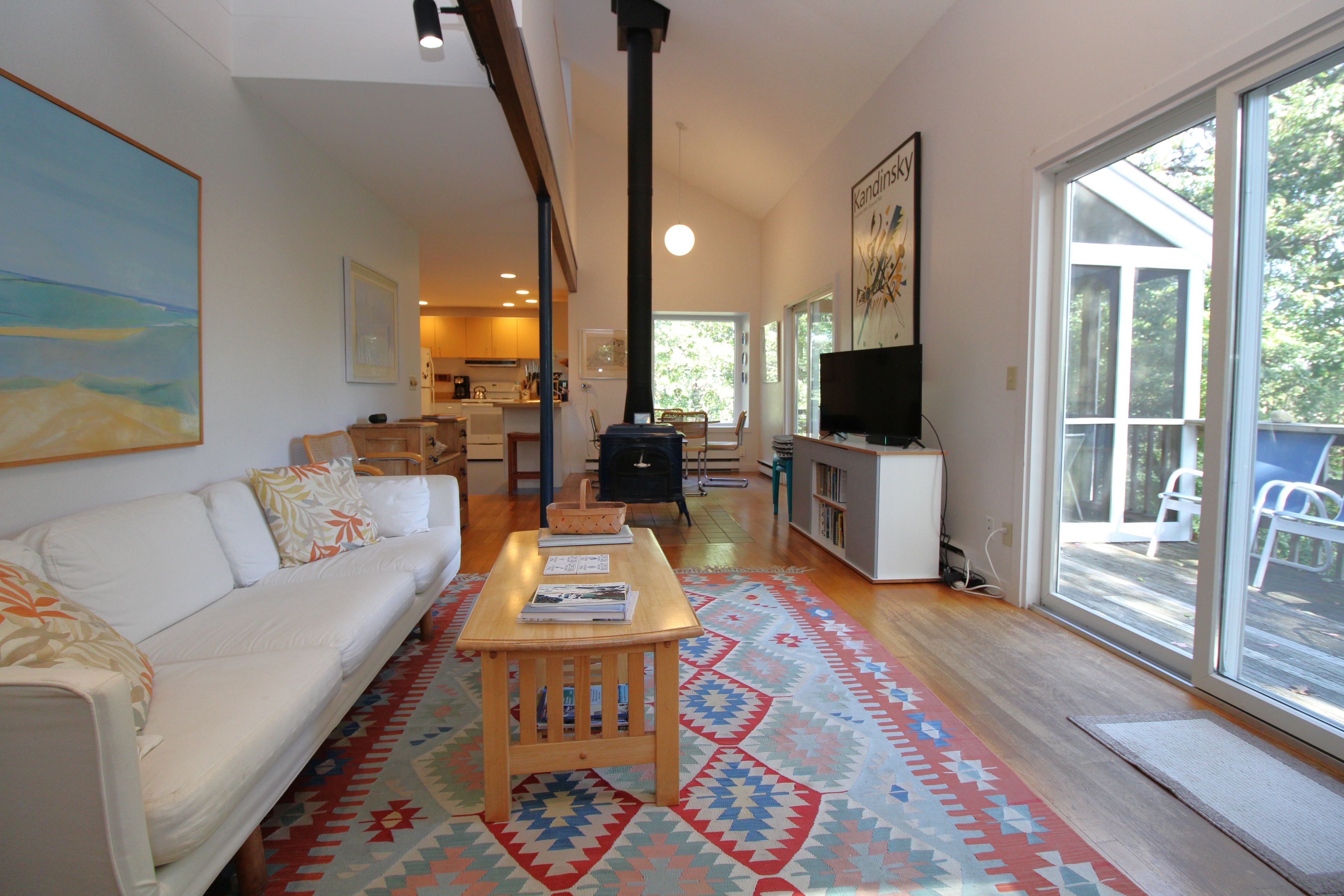 A living room with a sofa, coffee table, and a television. There is a kitchen in the background with cabinets and a refrigerator.