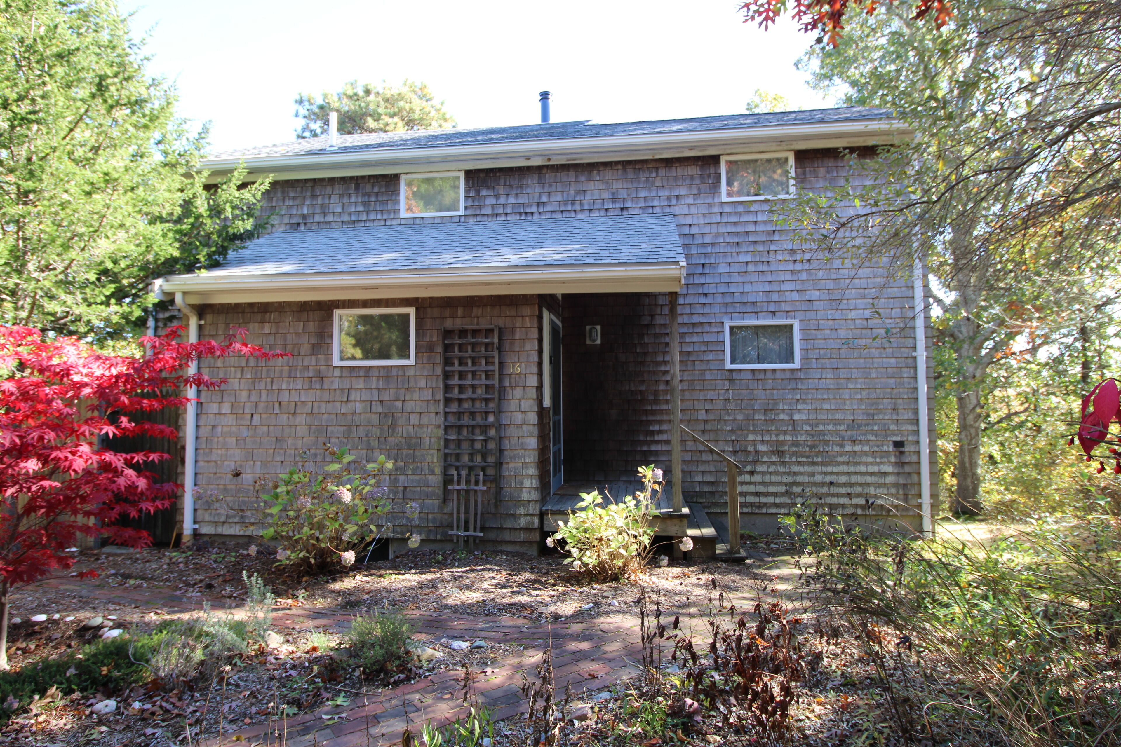A two-story house with a front porch, a garage door, and a small garden area with various plants and a red-leafed tree.