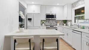 Una cocina moderna con armarios blancos, una isla central con dos sillas, un refrigerador de acero inoxidable, un microondas y un horno.
