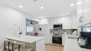 Una cocina moderna con armarios blancos, una isla central con taburetes y electrodomésticos como microondas, horno y cafetera.