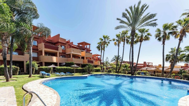 A swimming pool with a stone border, surrounded by palm trees and a building with balconies. - SR Ground floor flat with direct access to the pool! (Marbella)