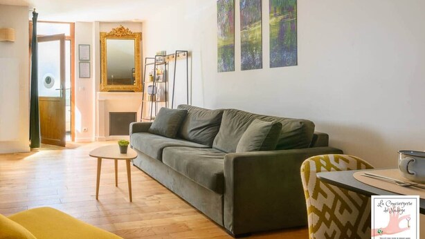 Een woonkamer met een bank, een klein tafeltje en een stoel. Er is een open haard, een spiegel en drie schilderijen aan de muur.