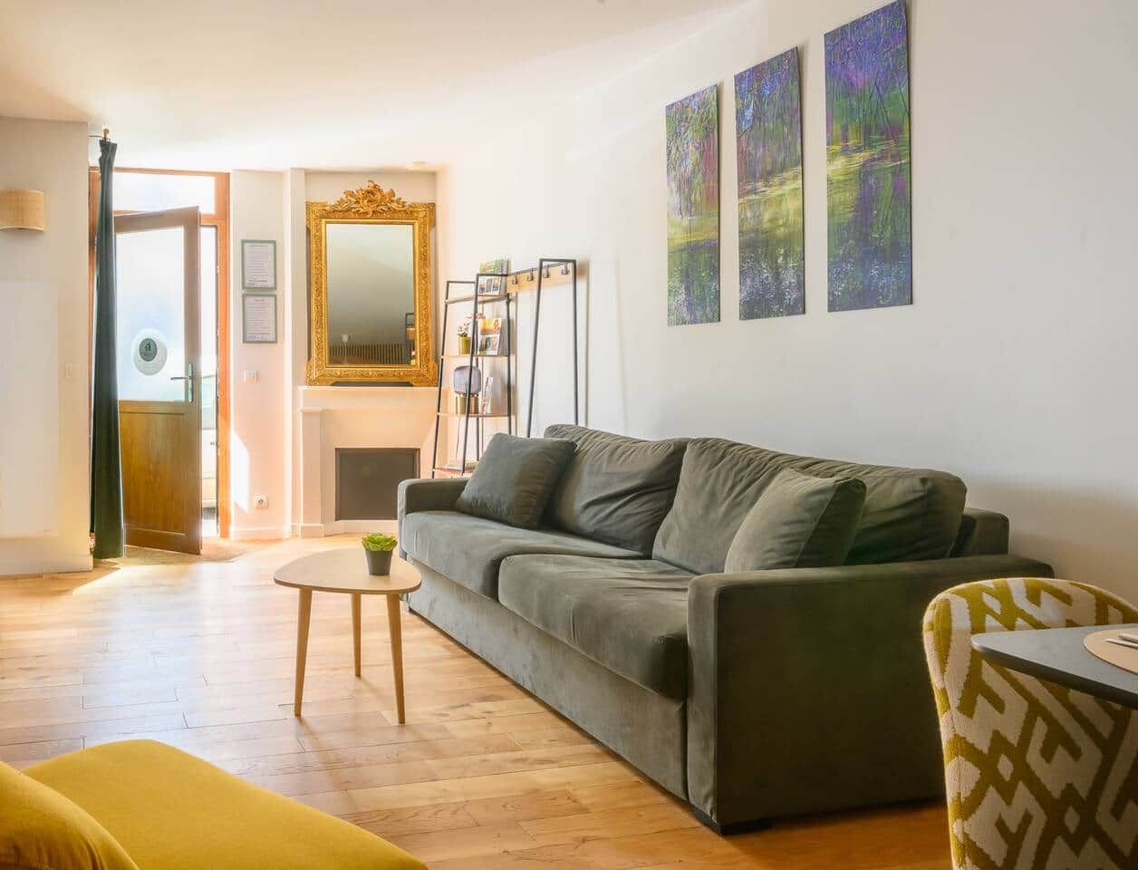 Un salon avec un canapé, une petite table et un fauteuil. On y trouve une cheminée, un miroir et trois tableaux accrochés au mur.