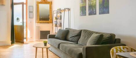 Un salon avec un canapé, une petite table et un fauteuil. On y trouve une cheminée, un miroir et trois tableaux accrochés au mur.