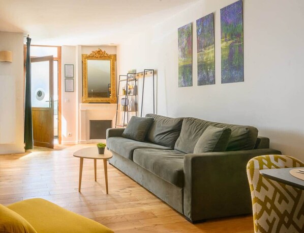A living room with a sofa, a small table, and a chair. There is a fireplace, a mirror, and three paintings on the wall.