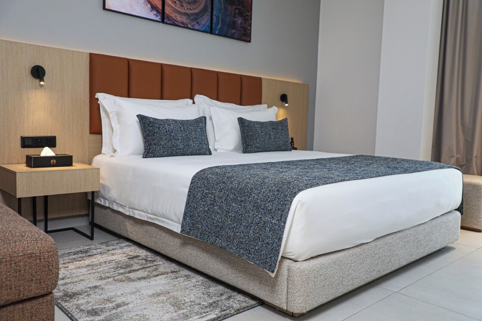 A modern hotel room with a large bed, a bedside table with a lamp, and a wall-mounted artwork.
